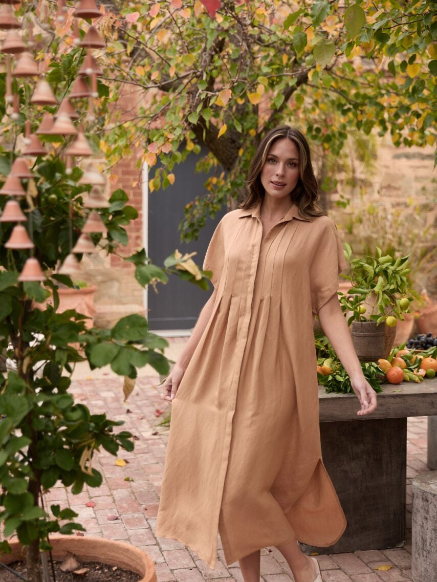 Woman in a beige dress standing in an outdoor setting with plants and a table.
